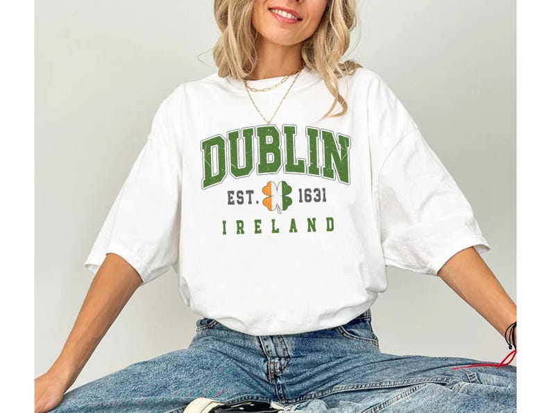Peut inclure: T-shirt blanc avec un tr&egrave;fle graphique vert, blanc et orange et le texte "DUBLIN EST. 1631 IRLANDE".