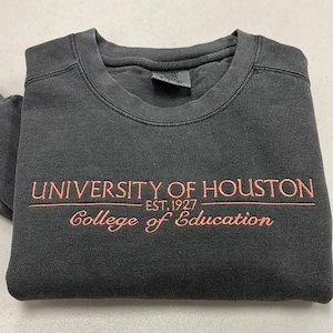 May include: A charcoal grey crewneck sweatshirt featuring the embroidered text UNIVERSITY OF HOUSTON EST. 1927 and College of Education in a light peach color. The garment has a ribbed collar and a faded, washed-out fabric appearance.