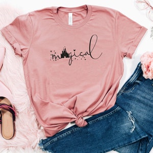 May include: A pink t-shirt with a black silhouette of Cinderella's castle and the word "magical" written in cursive.