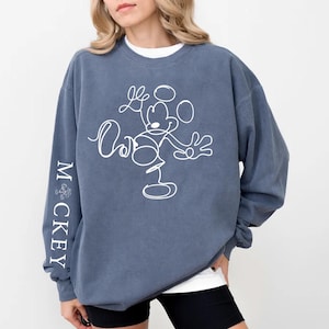 Puede incluir: Una sudadera azul con un contorno blanco de Mickey Mouse en la parte delantera y la palabra "MICKEY" escrita verticalmente en la manga izquierda.