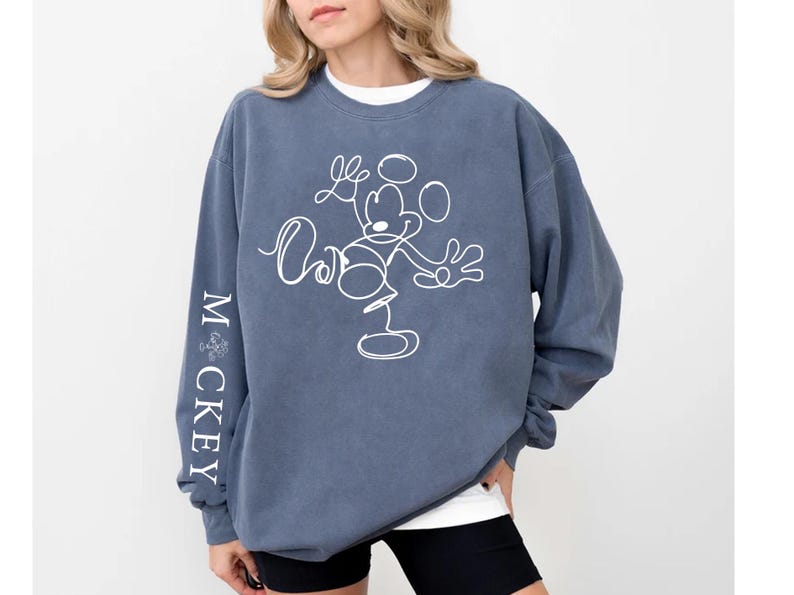Puede incluir: Una sudadera azul con un contorno blanco de Mickey Mouse en la parte delantera y la palabra "MICKEY" escrita verticalmente en la manga izquierda.