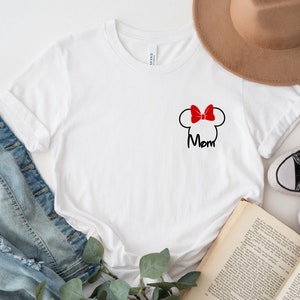 May include: White t-shirt with a black Minnie Mouse head outline, a red bow, and the word "Mom" in black script. The shirt is shown with a denim jacket, a brown hat, and black trainers.