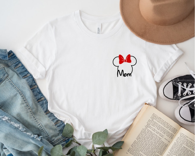 May include: White t-shirt with a black Minnie Mouse head outline, a red bow, and the word "Mom" in black script. The shirt is shown with a denim jacket, a brown hat, and black trainers.