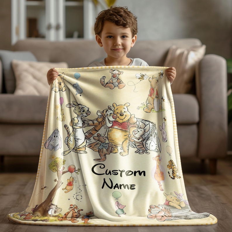 May include: A light yellow blanket featuring illustrations of Winnie the Pooh and friends. The blanket has a repeating pattern of characters and the text "Custom Name". The blanket is held by a child.