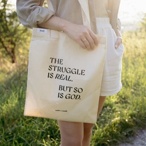 Könnte beinhalten: Eine weiße Canvas-Tragetasche mit schwarzem Text, der "The struggle is real. But so is God." lautet. Die Tasche wird von einer Person getragen, die ein hellbraunes Hemd und eine weiße Shorts trägt.