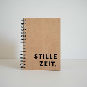 Könnte beinhalten: Ein braunes Spiralnotizbuch mit dem Text "STILLE ZEIT." in Schwarz auf dem Einband.