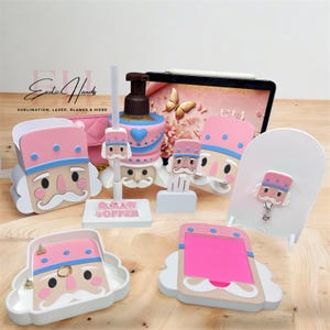 Puede incluir: Una colección de accesorios con temática de cascanueces en rosa, azul y beige. Los artículos incluyen un dispensador de jabón, un tapón para pajitas, un portabloc de notas, un portaanillos y un portainsignias. Los accesorios tienen un diseño de dibujos animados.