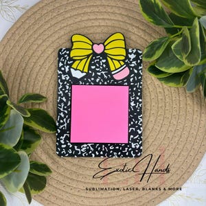 Puede incluir: Un portanotas con diseño de cuaderno de composición, con un lazo amarillo y un diseño de lápiz. Tiene un bloc de notas adhesivas rosa. El texto "Exotic Hands" y "SUBLIMATION, LASER, BLANKS & MORE" son visibles.