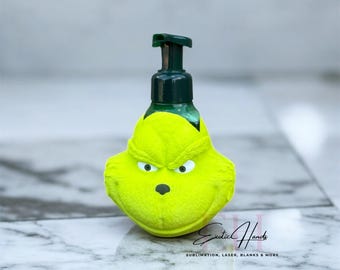 Puede incluir: Un dispensador de jabón verde con temática de Grinch con una bomba negra. El dispensador tiene una cara amarilla con ojos blancos y una nariz negra. El texto "Exotic Hands" está en la parte inferior.