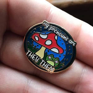 May include: Enamel pin with a red mushroom with white spots, a green frog, and the text "my pronouns are THEY THEM" on a blue background. The pin has a gold border.