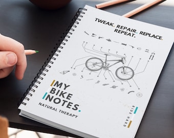 May include: A white spiral bound notebook with the text "MY BIKE INOTES. NATURAL THERAPY" and a black and white illustration of a bicycle with the text "TWEAK. REPAIR. REPLACE. REPEAT." and "RIDE HARD".