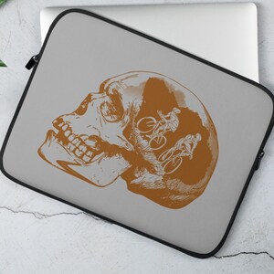 May include: A grey laptop sleeve featuring a brown skull graphic. The skull is outlined in black and has a design of two people riding bicycles inside it.