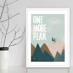 May include: A white framed print with a blue and green background featuring a mountain range and a silhouette of a person riding a bicycle over a peak. The text "ONE MORE PEAK" is in white letters on the print.