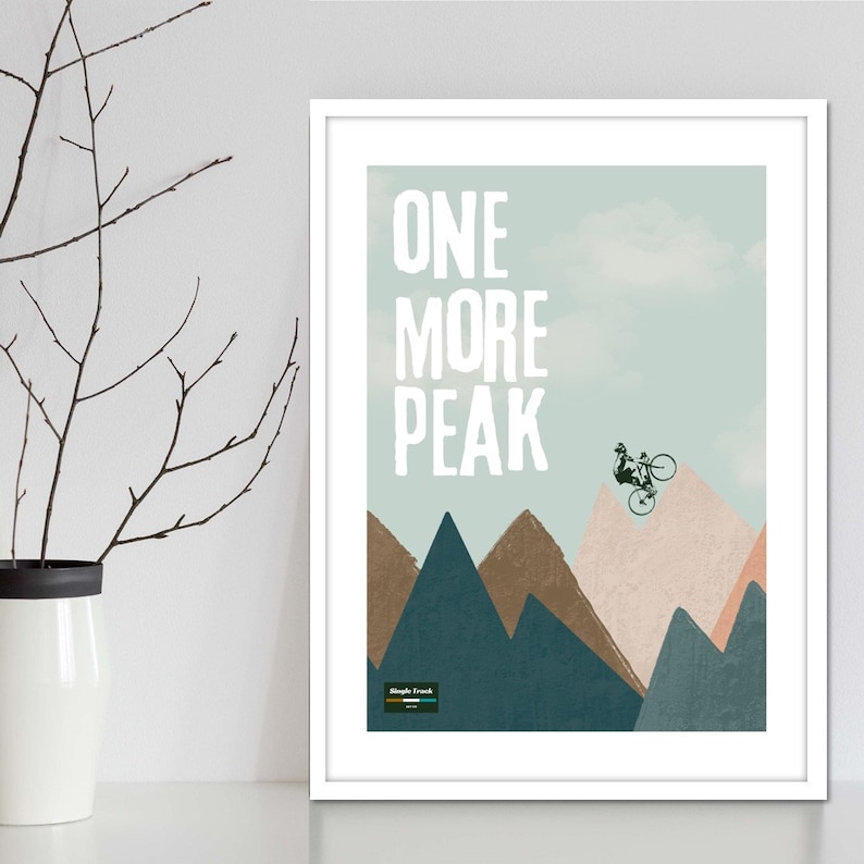 May include: A white framed print with a blue and green background featuring a mountain range and a silhouette of a person riding a bicycle over a peak. The text "ONE MORE PEAK" is in white letters on the print.