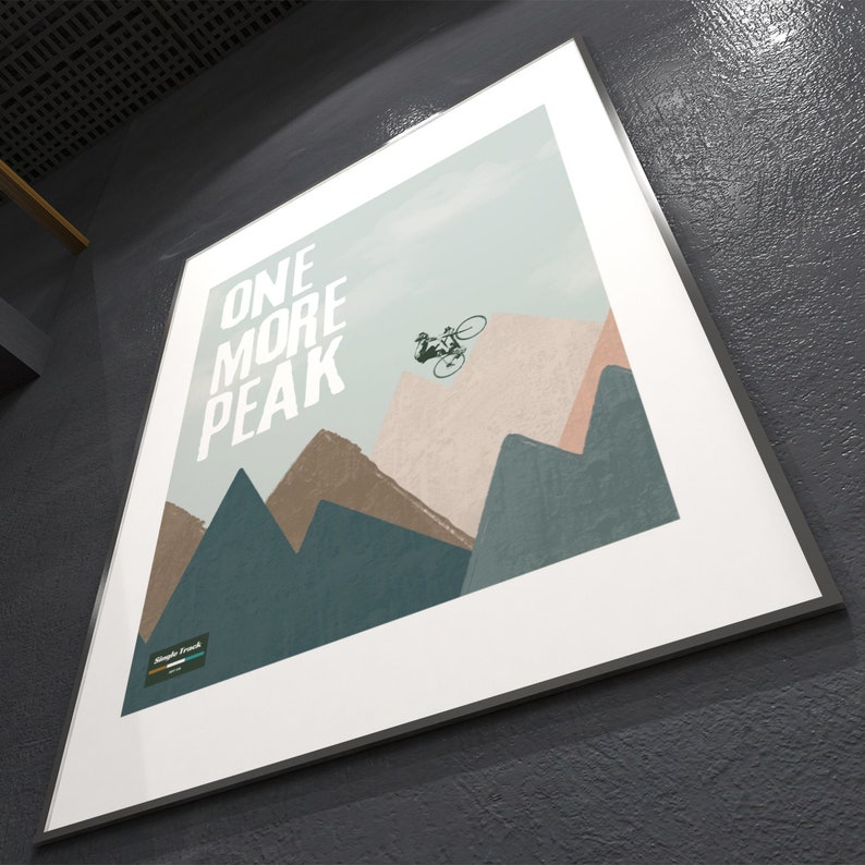 May include: A white framed poster with a blue, green, and brown mountain landscape. The poster features a silhouette of a person riding a bicycle over a mountain peak. The text "ONE MORE PEAK" is printed in white on the poster.