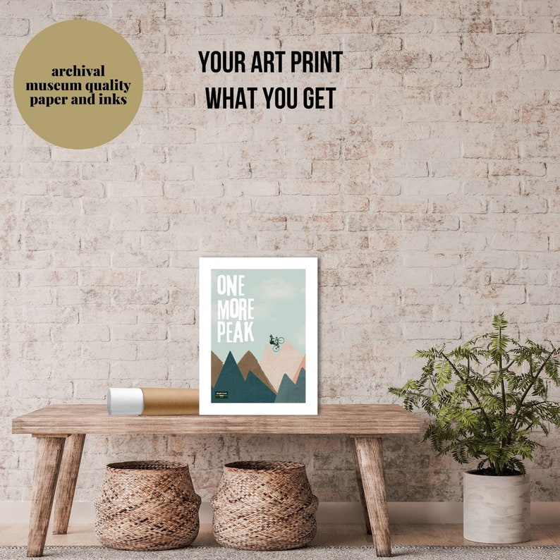 May include: A white framed art print with a mountain scene and a cyclist riding up a mountain. The text "ONE MORE PEAK" is printed in black on the print.