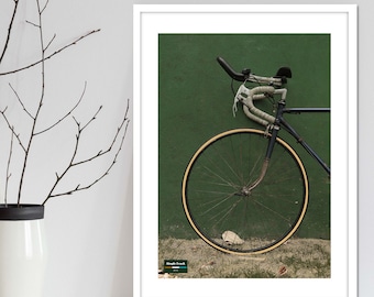 Op de afbeelding: Een vintage zwarte fiets met een wit stuur en witte tape op het stuur. De fiets leunt tegen een groene muur. De fiets heeft een bruine band met een witte velg. De fiets is in close-up.