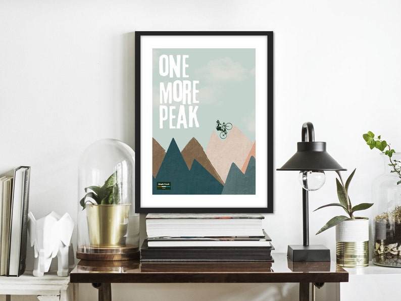 May include: A framed print with a mountain landscape and a cyclist jumping over a peak. The text "ONE MORE PEAK" is printed in white on a light blue background.