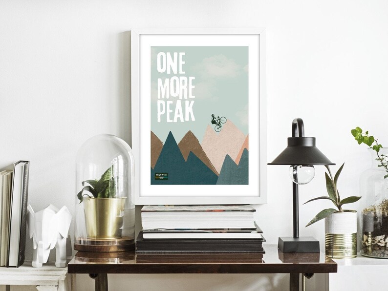 May include: A minimalist poster with a mountain range and a silhouette of a person riding a bicycle. The text "ONE MORE PEAK" is printed in white on a light blue background.
