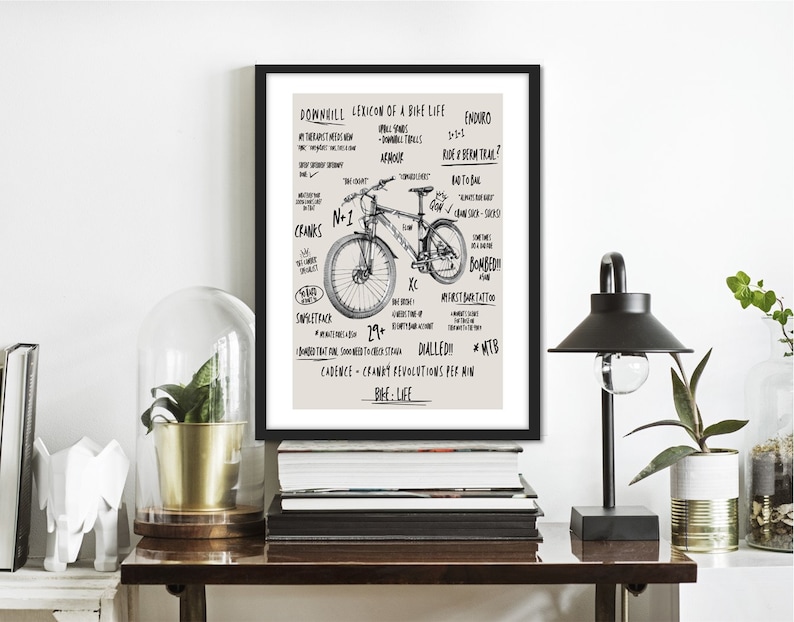 May include: A black and white illustration of a mountain bike with the text "Downhill Lexicon of a Bike Life" and other related terms. The illustration is framed in a black frame and is hanging on a white wall.