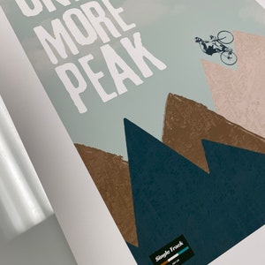 May include: A white graphic print with the text "ONE MORE PEAK" on a blue and brown mountain background. A silhouette of a person riding a bicycle is in the upper right corner. The text "Single Track" is in a black rectangle with a blue and green stripe.