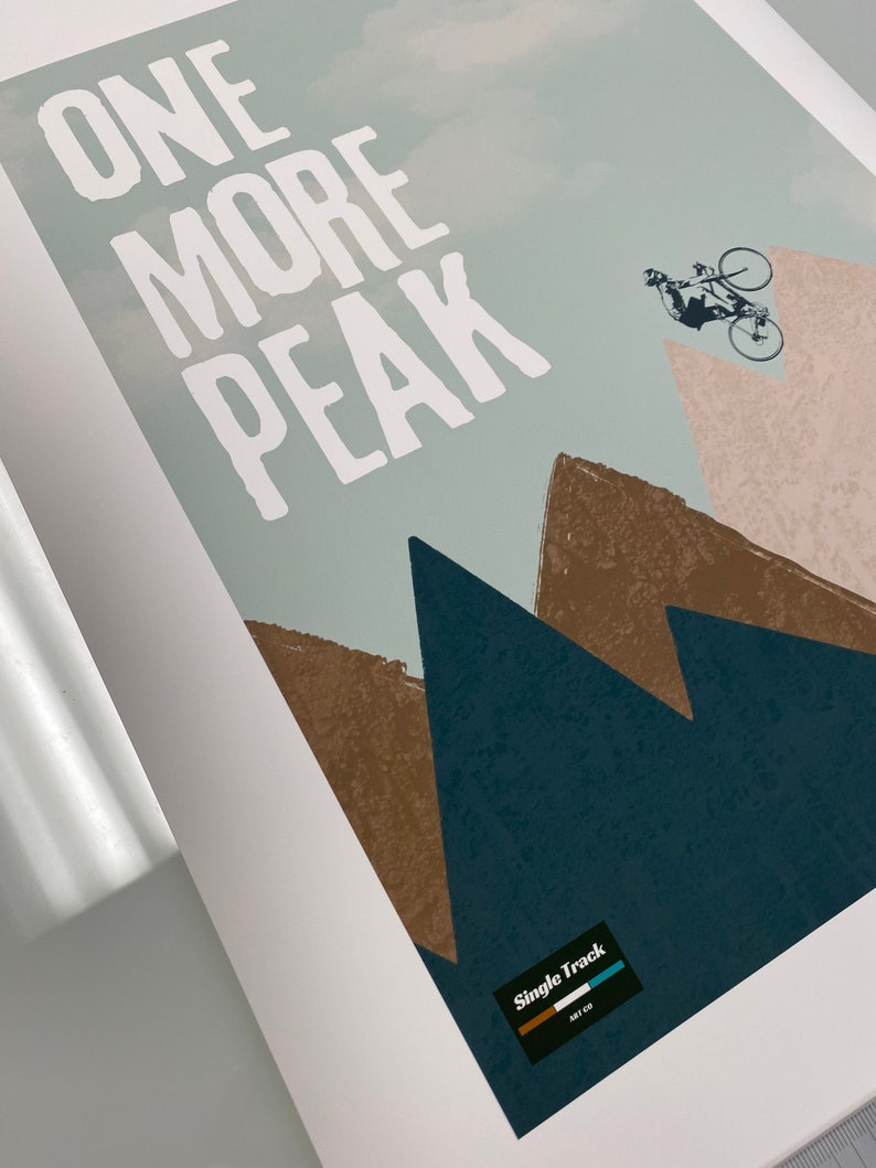 May include: A white graphic print with the text "ONE MORE PEAK" on a blue and brown mountain background. A silhouette of a person riding a bicycle is in the upper right corner. The text "Single Track" is in a black rectangle with a blue and green stripe.