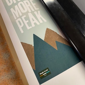 May include: A white print with the text "ONE MORE PEAK" and a graphic of three brown mountains on a blue background. The print has a logo in the bottom right corner that says "Single Track" with a small logo of a mountain range.