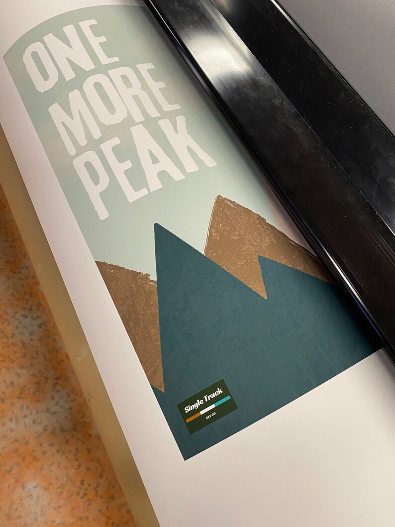 May include: A white print with the text "ONE MORE PEAK" and a graphic of three brown mountains on a blue background. The print has a logo in the bottom right corner that says "Single Track" with a small logo of a mountain range.