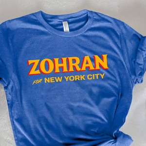May include: A blue t-shirt with the text "ZOHRAN for NEW YORK CITY" in yellow and orange lettering. The shirt is knotted at the bottom. A straw hat and a striped scarf are visible in the background.