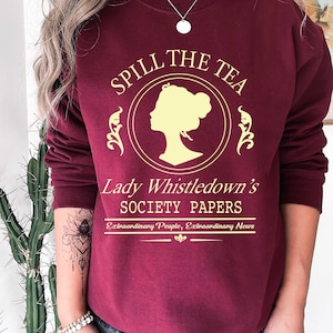 May include: Burgundy sweatshirt with the text "Spill the Tea" in gold lettering, featuring a silhouette of a woman's profile. Below the text is "Lady Whistledown's Society Papers" and "Extraordinary People, Extraordinary News".
