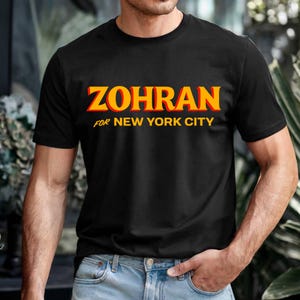 May include: Black t-shirt with the word "ZOHRAN" in orange and yellow block letters. Below the word "ZOHRAN" is the text "FOR NEW YORK CITY" in yellow. The t-shirt is being worn.