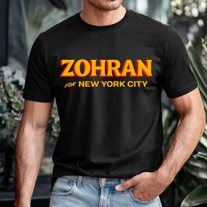 May include: Black t-shirt with the word "ZOHRAN" in orange and yellow block letters. Below the word "ZOHRAN" is the text "FOR NEW YORK CITY" in yellow. The t-shirt is being worn.