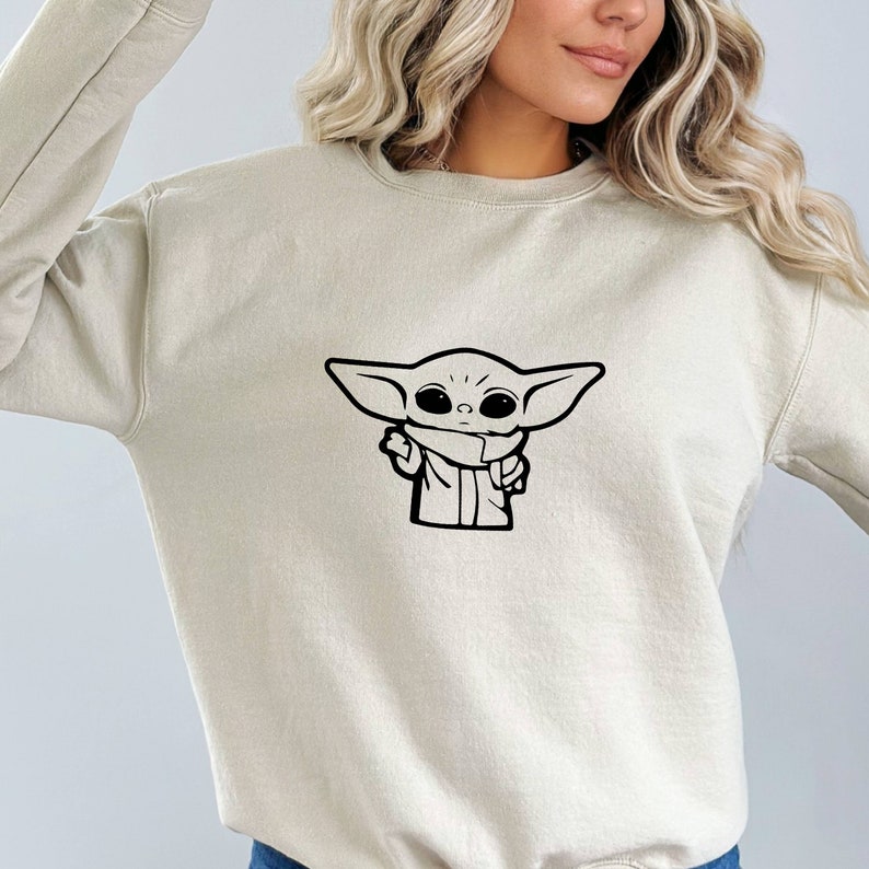 May include: A light beige sweatshirt with a black and white illustration of a character from the Star Wars franchise, The Mandalorian, known as "The Child" or "Baby Yoda".