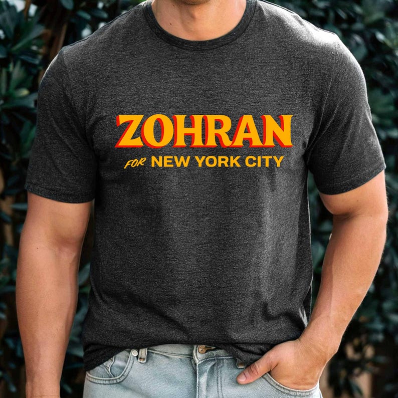May include: Dark gray t-shirt with the word "ZOHRAN" in yellow and red block letters. Below the word is the phrase "FOR NEW YORK CITY" in a smaller, yellow font. The shirt is a crew neck style.