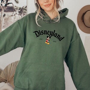 May include: Green hooded sweatshirt with a black Disneyland logo and Mickey Mouse graphic. The logo reads "Disneyland" with "EST. 1955" underneath.