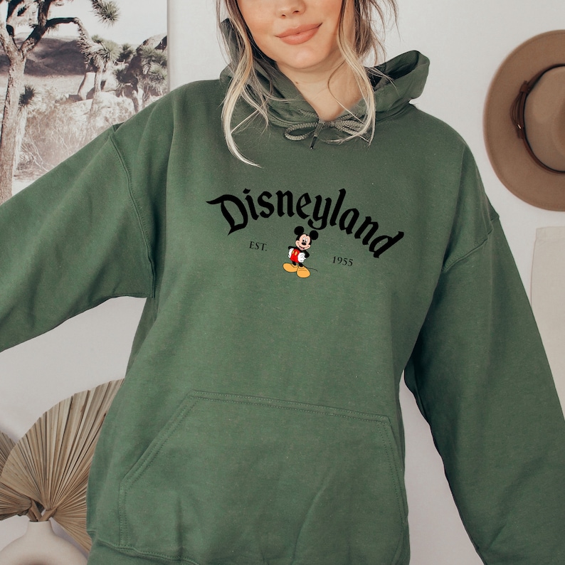 May include: Green hooded sweatshirt with a black Disneyland logo and Mickey Mouse graphic. The logo reads "Disneyland" with "EST. 1955" underneath.