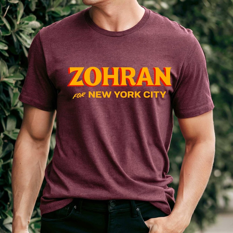 May include: Maroon crew neck t-shirt with the text "ZOHRAN" in yellow with a red outline, and the text "FOR NEW YORK CITY" in yellow. The shirt is made of a soft material.