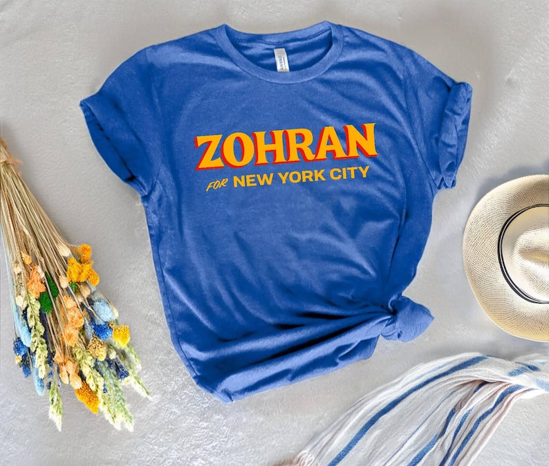 May include: A blue t-shirt with the text "ZOHRAN for NEW YORK CITY" in yellow and orange lettering. The shirt is knotted at the bottom. A straw hat and a striped scarf are visible in the background.