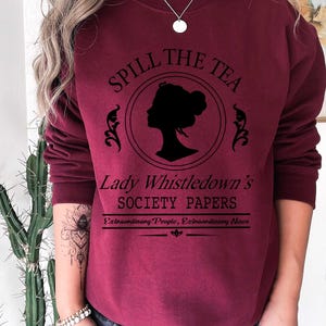 May include: Burgundy sweatshirt with the text "Spill the Tea" above a silhouette of a woman's profile in a circle. Below the circle is "Lady Whistledown's Society Papers" and "Extraordinary People, Extraordinary News".