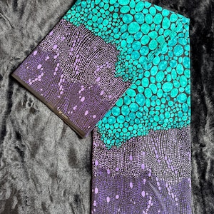 Puede incluir: Tela doblada con un patrón vibrante. La tela presenta un diseño turquesa y negro de formas circulares, y un patrón de puntos morados y negros. La palabra "ORIGINAL" está impresa en la tela.