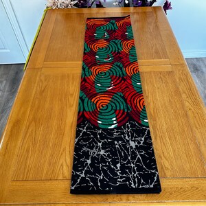 May include: A rectangular table runner with a vibrant spiral pattern in red, green, and orange on a black background. The bottom section features a black and white abstract design. The runner is placed on a wooden table.