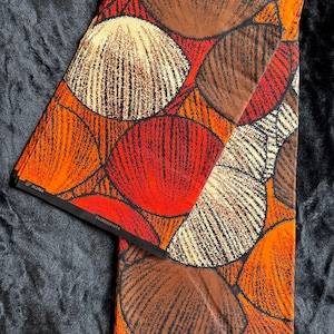 Könnte beinhalten: Gefalteter Stoff mit einem lebendigen Muster aus großen, stilisierten Muschelformen in Orange-, Rot-, Braun- und Cremetönen, umrandet in Schwarz. Der Stoff hat ein glänzendes Finish und das Wort "FASHIONISTA" ist sichtbar.