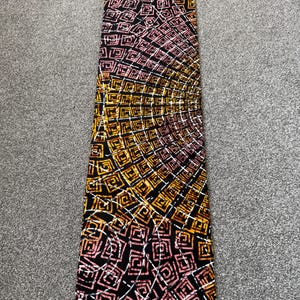 May include: A rectangular fabric with a geometric pattern in black, yellow, and pink. The design features squares and lines arranged in a radial pattern. The fabric is likely used for clothing or home decor.