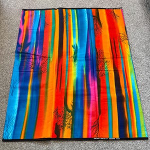 May include: A colourful fabric featuring vertical stripes in blue, green, yellow, orange, and red. Black tree silhouettes are scattered across the vibrant background. The fabric has a black border with printed text.