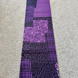 May include: A rectangular piece of fabric with a patchwork design in black and purple. The fabric features various patterns, including geometric shapes, lines, and squiggles. The text "GUARANTEED DUTCH WAX HOLLAND" is printed at the bottom.
