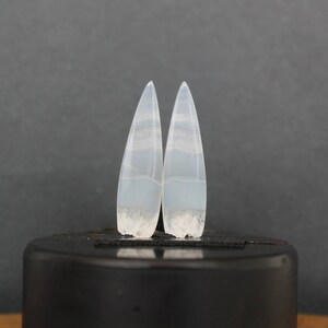 Puede incluir: Par de cabujones de ágata blanca con forma puntiaguda y patrón en capas. Los cabujones están colocados sobre una superficie negra.