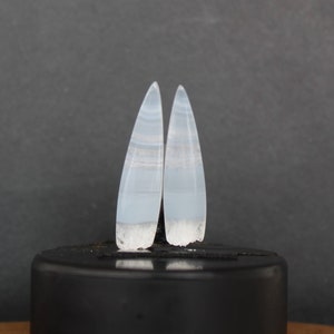 Puede incluir: Un par de pendientes de ágata azul claro con forma puntiaguda de lágrima. Los pendientes tienen un patrón blanco a rayas y un acabado esmerilado y texturizado.