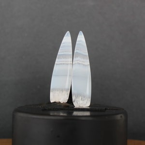 Puede incluir: Dos piedras preciosas pulidas y alargadas con un patrón de bandas azul claro y blanco. Las piedras tienen un acabado liso y brillante y una base rugosa y texturizada. Se alzan sobre un fondo oscuro, mostrando su belleza natural y diseño único.