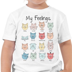 May include: White t-shirt with the text "My Feelings" and a graphic of 16 cartoon cats with different facial expressions representing various emotions. Each cat has a word underneath it describing the emotion, such as happy, sad, angry, and scared.