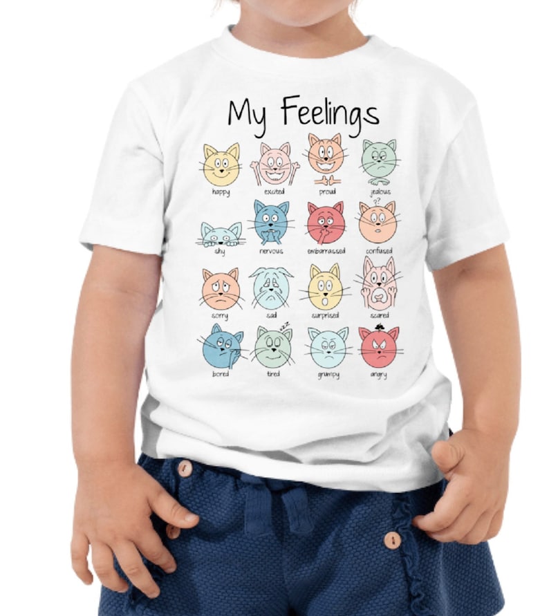 May include: White t-shirt with the text "My Feelings" and a graphic of 16 cartoon cats with different facial expressions representing various emotions. Each cat has a word underneath it describing the emotion, such as happy, sad, angry, and scared.